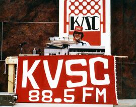 Man sits at a KVSC radio booth in front of Stewart Hall (1948), St. Cloud State University